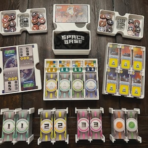 May include: A board game set with cards, tokens, dice, and player boards. The game includes a white box with the text "SPACE BASE" on a black bag. The game pieces are arranged in white trays. The game components are on a dark wooden surface.