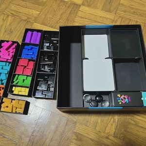 Puede incluir: Una caja de juego de mesa abierta con varios componentes. La caja contiene formas geom&eacute;tricas coloridas en rosa, azul, naranja, morado, rojo, verde y amarillo. Tambi&eacute;n hay piezas de juego negras, cartas blancas y esferas negras. La caja del juego es negra con un acento turquesa.