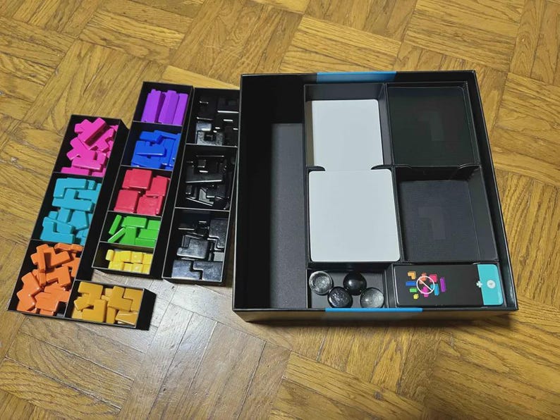Puede incluir: Una caja de juego de mesa abierta con varios componentes. La caja contiene formas geom&eacute;tricas coloridas en rosa, azul, naranja, morado, rojo, verde y amarillo. Tambi&eacute;n hay piezas de juego negras, cartas blancas y esferas negras. La caja del juego es negra con un acento turquesa.