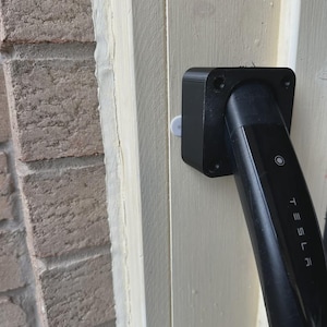 以下が含まれることがあります： 明るい色の木材の表面に取り付けられた黒いテスラ充電コネクタ。コネクタは長方形で、端が丸みを帯びており、テスラのロゴが見えます。表面はレンガの壁の隣にあります。