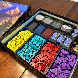 Puede incluir: Caja de juego de mesa abierta con compartimentos llenos de piezas y cartas de colores. La caja contiene piezas de madera amarillas, turquesas, rojas y moradas. La tapa de la caja del juego muestra el título "Imperial Steam".