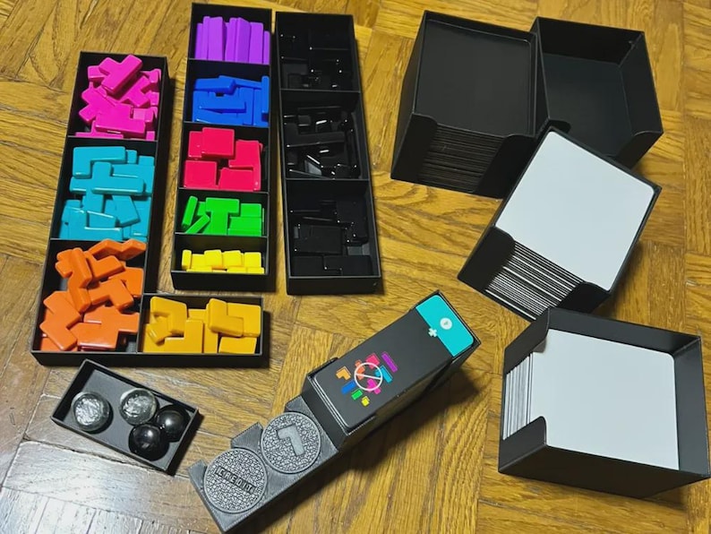 Puede incluir: Un juego de mesa con formas geom&eacute;tricas coloridas en bandejas negras. El juego incluye varios bloques de colores, cartas y fichas. Las piezas del juego son en tonos de rosa, azul, naranja, amarillo, verde y morado. El juego est&aacute; sobre una superficie de madera.