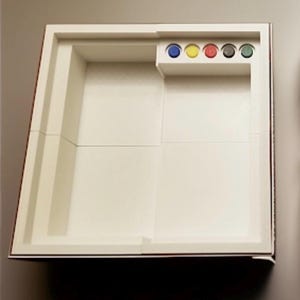 Pu&ograve; includere: Un tabellone di gioco rettangolare bianco con un'area di gioco incassata e un piccolo scomparto contenente pedine colorate. Il tabellone ha un bordo rosso ed &egrave; progettato per un gioco da tavolo.