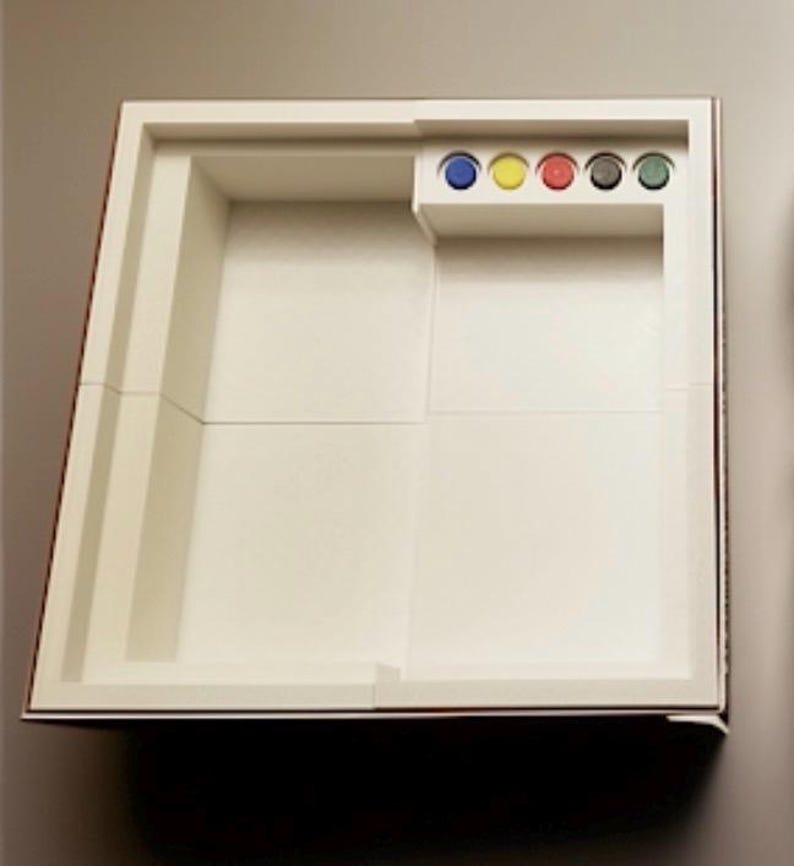 Pu&ograve; includere: Un tabellone di gioco rettangolare bianco con un'area di gioco incassata e un piccolo scomparto contenente pedine colorate. Il tabellone ha un bordo rosso ed &egrave; progettato per un gioco da tavolo.