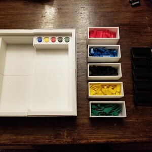 Pu&ograve; includere: Un organizer per giochi da tavolo bianco con scomparti per i pezzi del gioco. Include piccole scatole bianche piene di pezzi del gioco rossi, blu, neri, gialli e verdi. Include anche una pila di porta carte neri e carte da gioco.