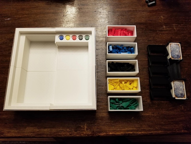 Pu&ograve; includere: Un organizer per giochi da tavolo bianco con scomparti per i pezzi del gioco. Include piccole scatole bianche piene di pezzi del gioco rossi, blu, neri, gialli e verdi. Include anche una pila di porta carte neri e carte da gioco.