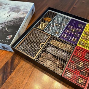 May include: A board game box set titled "Brass: Birmingham" with a detailed cityscape illustration. The open box reveals various game components in colorful, honeycomb-patterned trays, including yellow, red, and purple.