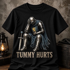 Op de afbeelding: Een zwart t-shirt met een afbeelding van een ridder in zilveren harnas die zit met één hand op het hoofd en de andere op de buik. De ridder draagt een blauwe cape met een geel leeuwembleem en houdt een zwaard vast. De tekst "TUMMY HURTS" staat eronder.
