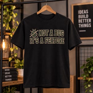 May include: A black short-sleeved t-shirt featuring a graphic of a beetle with a diagonal line through it, accompanied by the text "NOT A BUG IT'S A FEATURE" in a bold, collegiate-style font with light green lettering and dark green outlines.