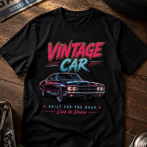 May include: A black short-sleeved t-shirt featuring a graphic print of a classic car. The text reads VINTAGE CAR in pink and blue, with BUILT FOR THE ROAD and Live to Drive printed below the car illustration in pink and teal lettering.