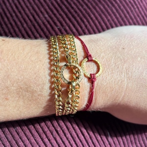 May include: A gold-toned multi-strand chain bracelet featuring a central circular link with a textured pattern, paired with a thin red cord bracelet that includes a matching gold-toned circular link with a textured surface design.