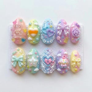 May include: A set of ten pastel-colored, decorated artificial nails. Each nail features a different design, including bows, hearts, stars, and a teddy bear. The nails are arranged on a white surface, showcasing a variety of colors and embellishments.