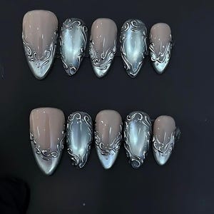 May include: A set of ten almond-shaped false nails. The nails alternate between a nude pink and a metallic silver-gray. The silver-gray nails feature an ornate, swirling design along the base.