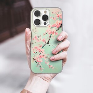 Peut inclure: Une coque de téléphone transparente avec un motif floral représentant des fleurs de cerisier roses sur un fond vert clair. La coque est conçue pour un iPhone avec un renflement de l'appareil photo.