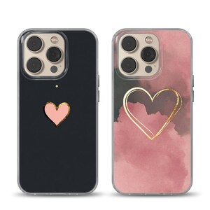 May include: Two phone cases with heart designs. One case is black with a pink heart and gold outline. The other case has a pink and gray watercolor background with a gold heart outline. Both cases have a clear border.