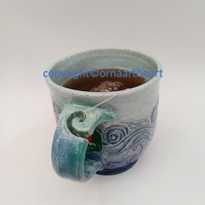 Op de afbeelding: Een blauwe en witte keramische mok met een golfontwerp. De mok is gevuld met thee en heeft een theezakje dat aan de rand hangt.