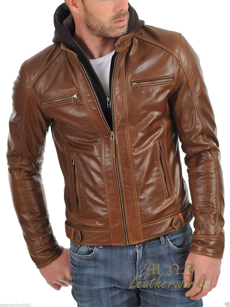 May include: Brown leather jacket with a zip-up front, multiple zippered pockets, and a hooded lining. The jacket has a classic design with a tailored fit. The model is wearing blue jeans. The jacket is from M.N. Leatherworks.