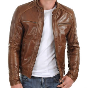 May include: A brown leather jacket with a stand-up collar and multiple zippered pockets. The jacket has a classic design and a slightly distressed appearance. The model is wearing a white t-shirt and blue jeans.