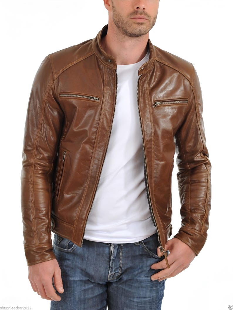 May include: A brown leather jacket with a stand-up collar and multiple zippered pockets. The jacket has a classic design and a slightly distressed appearance. The model is wearing a white t-shirt and blue jeans.