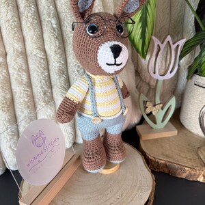 Puede incluir: Muñeca de conejo de ganchillo con gafas, camiseta a rayas amarillas y blancas, pantalones cortos azules y tirantes. El conejo marrón tiene detalles grises y está sobre una rodaja de madera. Un letrero transparente con el texto "Winning Stitches" está al frente.