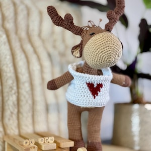 Könnte beinhalten: Ein brauner gehäkelter Elch, der ein weißes gehäkeltes Hemd mit einem roten Herzen auf der Vorderseite trägt. Der Elch steht auf einem Holzschlitten mit den Worten "PATCH" und "PA" an der Seite.