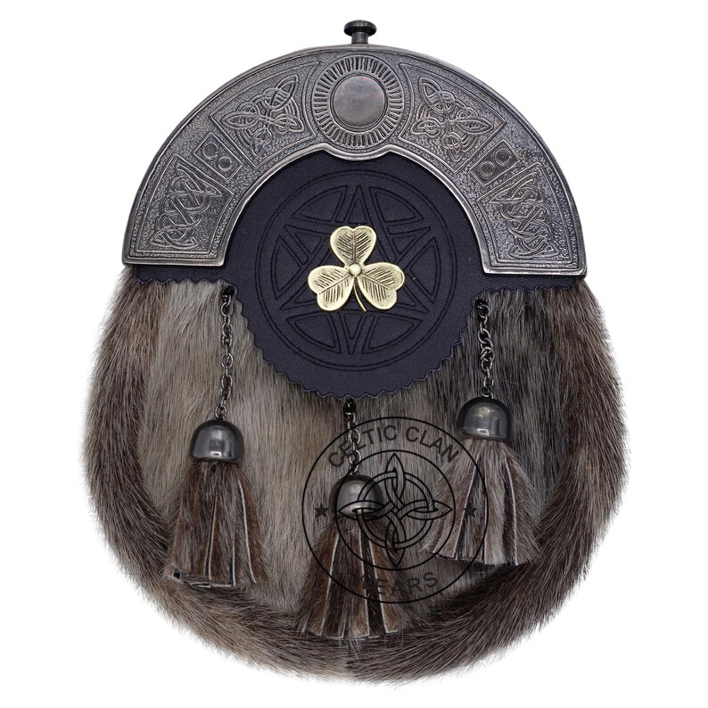 K&ouml;nnte beinhalten: Ein traditioneller schottischer Kilt-Sporran mit einem Fellk&ouml;rper und einem dekorativen Metall-Cantle. Der Cantle zeigt keltische Knotenmuster und ein zentrales Emblem. Zwei Quasten h&auml;ngen darunter, und ein goldenes Kleeblatt ist auf der Lederklappe zentriert.
