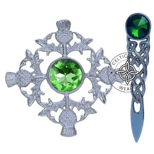 K&ouml;nnte beinhalten: Zwei silberne Broschen mit gr&uuml;nen Kristall-Akzenten. Eine Brosche zeigt ein keltisches Knotendesign und die andere ein Kleeblatt-Design mit Distel-Akzenten.