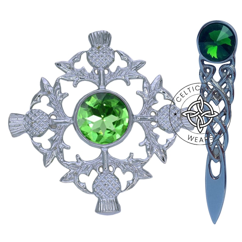 K&ouml;nnte beinhalten: Zwei silberne Broschen mit gr&uuml;nen Kristall-Akzenten. Eine Brosche zeigt ein keltisches Knotendesign und die andere ein Kleeblatt-Design mit Distel-Akzenten.