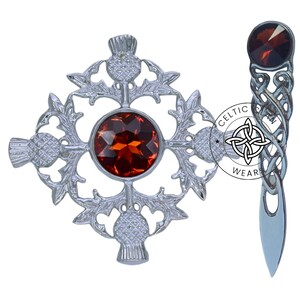 K&ouml;nnte beinhalten: Eine silberne Distelbrosche mit einem gro&szlig;en orangefarbenen Edelstein in der Mitte. Die Brosche ist von vier kleineren Disteln umgeben. Die Brosche befindet sich auf einem wei&szlig;en Hintergrund. Celtic Wear.