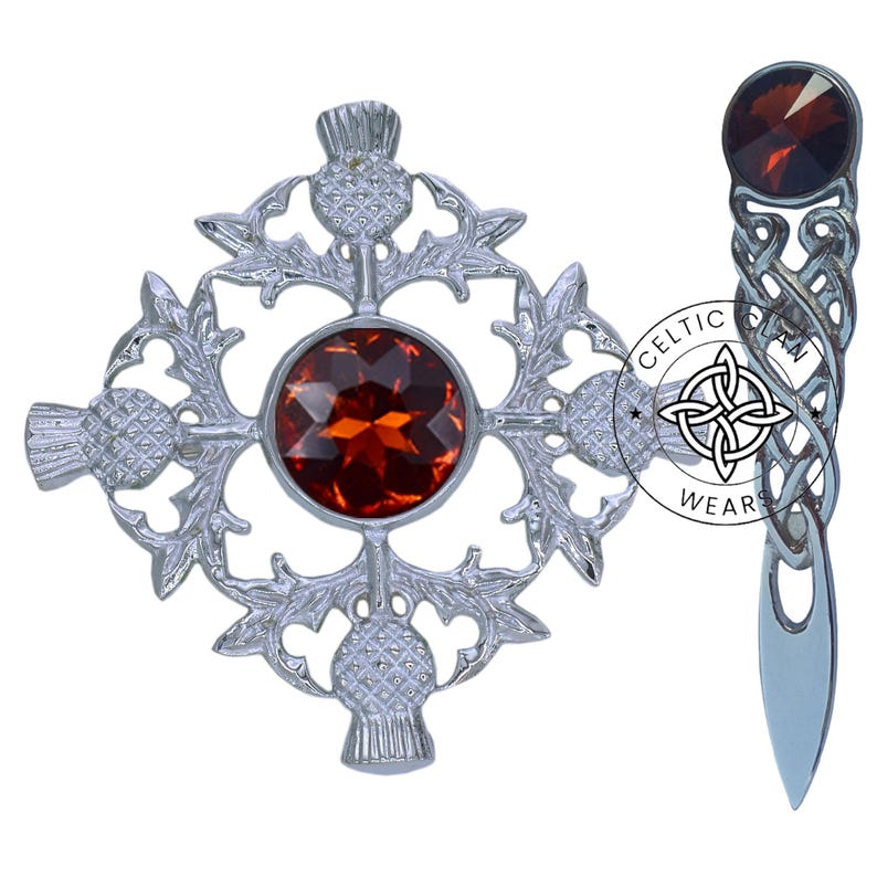 K&ouml;nnte beinhalten: Eine silberne Distelbrosche mit einem gro&szlig;en orangefarbenen Edelstein in der Mitte. Die Brosche ist von vier kleineren Disteln umgeben. Die Brosche befindet sich auf einem wei&szlig;en Hintergrund. Celtic Wear.