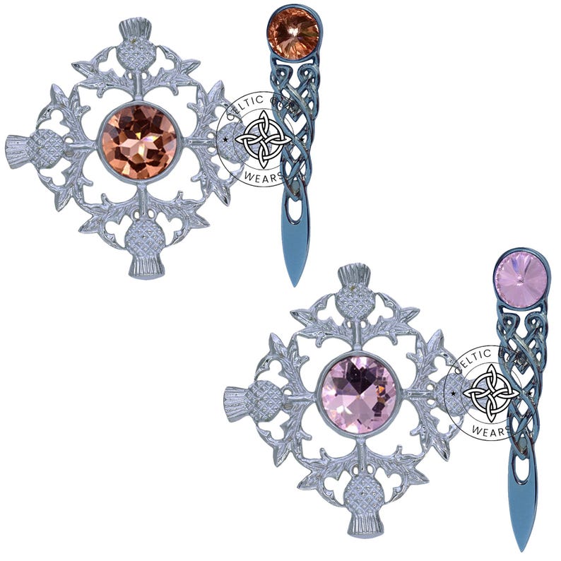 K&ouml;nnte beinhalten: Zwei silberne Kiltnadeln mit keltischen Knoten, Distelmotiven und Kristallverzierungen. Eine Nadel hat einen rosa Kristall, die andere einen orangefarbenen Kristall.