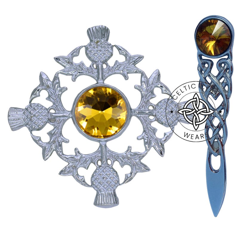K&ouml;nnte beinhalten: Ein Set aus zwei silberfarbenen Broschen mit einem keltischen Knotendesign und einem Distel-Design mit einem gelben Kristall. Die Distelbrosche hat vier Disteln und einen gro&szlig;en gelben Kristall in der Mitte. Die keltische Knotenbrosche hat einen einzelnen gelben Kristall oben. Der Text "Celtic Wear" ist auf der Brosche aufgedruckt.
