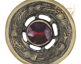 Könnte beinhalten: Runde Brosche im antiken Stil mit einem zentralen roten Edelstein. Die Brosche hat einen bronzefarbenen Metallrahmen mit einem detaillierten Disteldesign am Rand. Das Wort "Red" ist oben links zu sehen.