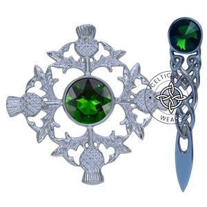 K&ouml;nnte beinhalten: Eine silberne Brosche mit einem keltischen Knoten und einem gr&uuml;nen Stein sowie eine silberne Distelbrosche mit einem gr&uuml;nen Stein. Die Distelbrosche hat vier Disteln und ein keltisches Knotendesign.