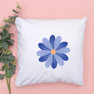 May include: White square pillow featuring a large blue daisy design with a yellow center. The daisy has multiple petals in varying shades of blue. The pillow is on a pink background with green foliage.