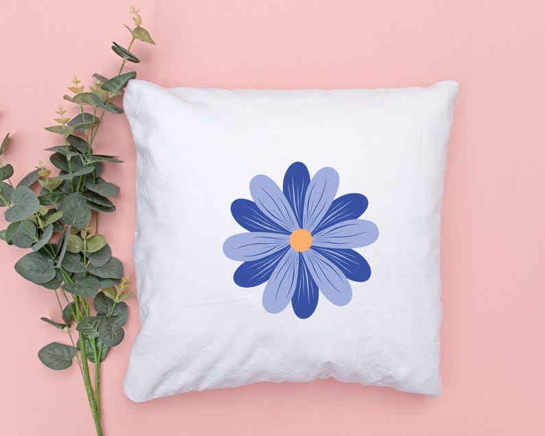 May include: White square pillow featuring a large blue daisy design with a yellow center. The daisy has multiple petals in varying shades of blue. The pillow is on a pink background with green foliage.