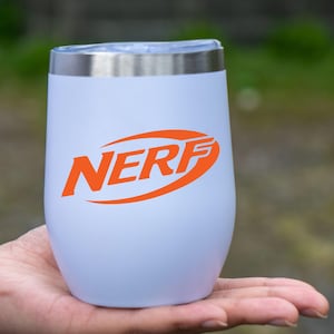 May include: A white insulated tumbler with a silver rim and lid. The orange "NERF" logo is prominently displayed on the front. The tumbler is held in a person's hand against a blurred green background.
