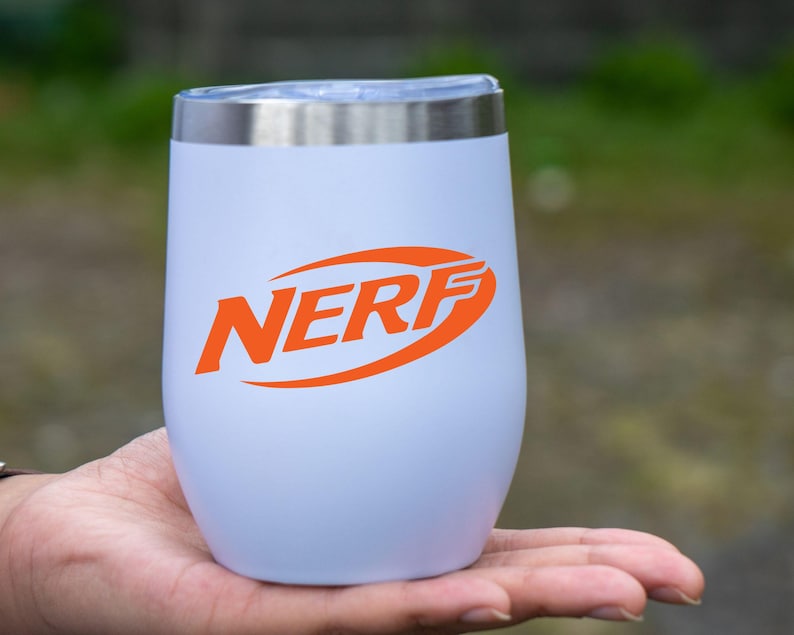 May include: A white insulated tumbler with a silver rim and lid. The orange "NERF" logo is prominently displayed on the front. The tumbler is held in a person's hand against a blurred green background.