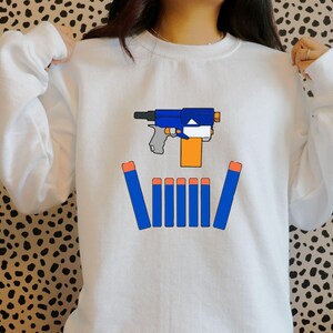 May include: White sweatshirt featuring a graphic of a blue and orange toy gun with blue darts. The design is centered on the front of the sweatshirt. The sweatshirt has long sleeves and a crew neck.