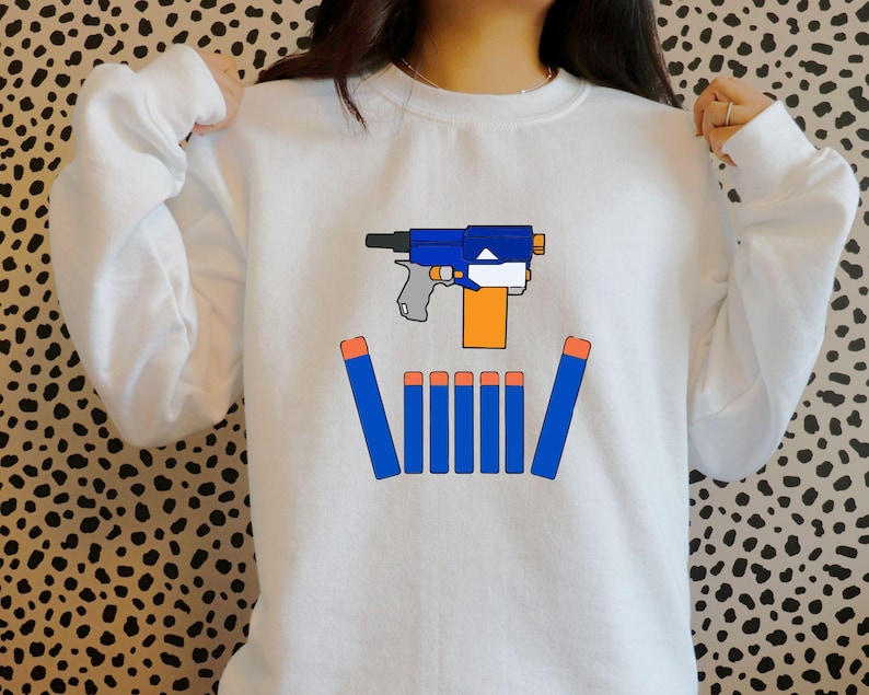 May include: White sweatshirt featuring a graphic of a blue and orange toy gun with blue darts. The design is centered on the front of the sweatshirt. The sweatshirt has long sleeves and a crew neck.