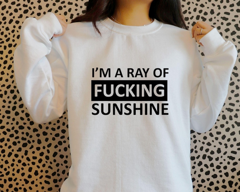 May include: White sweatshirt with the text "I'M A RAY OF FUCKING SUNSHINE" in black block letters. The text is centered within a black rectangle. The sweatshirt has long sleeves and a crew neck.