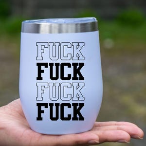 May include: A white insulated tumbler with a silver rim, featuring the word "FUCK" stacked four times in a bold, black, collegiate-style font. The tumbler is held in a hand against a blurred green background.