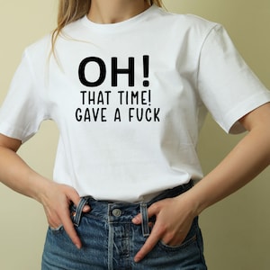 May include: White t-shirt with the text "OH! THAT TIME! GAVE A FUCK" in black. The shirt is being worn with blue jeans. The person is standing in front of a light beige background.