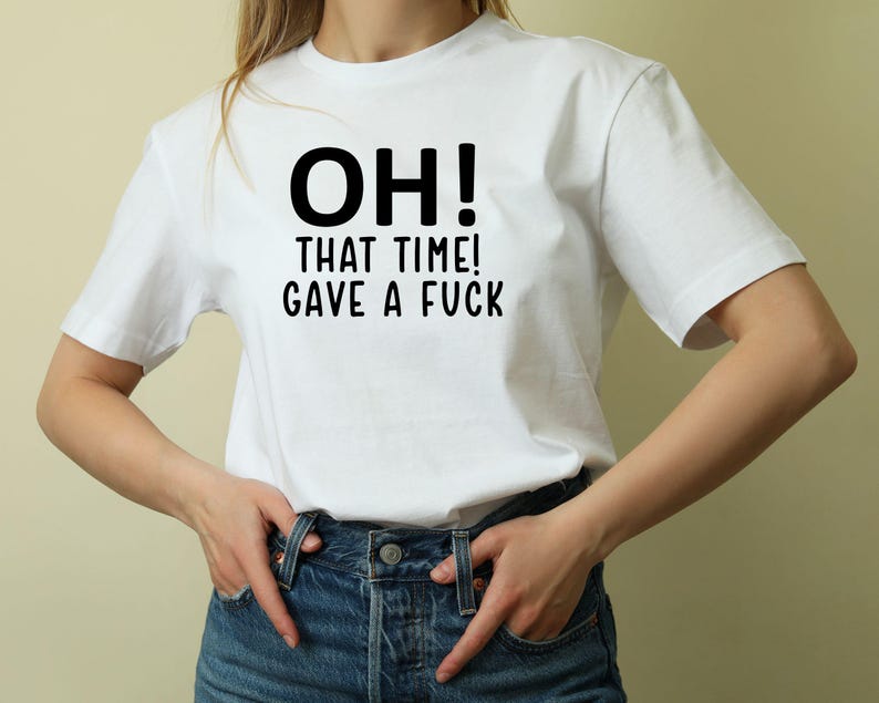 May include: White t-shirt with the text "OH! THAT TIME! GAVE A FUCK" in black. The shirt is being worn with blue jeans. The person is standing in front of a light beige background.