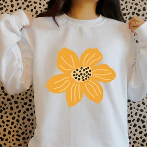 May include: White sweatshirt featuring a large, stylized yellow flower graphic on the front. The flower has a black dotted center and curved petal lines. The sweatshirt has long sleeves and a crew neck.