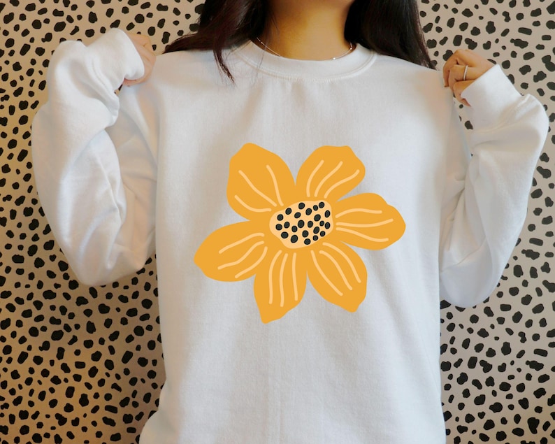 May include: White sweatshirt featuring a large, stylized yellow flower graphic on the front. The flower has a black dotted center and curved petal lines. The sweatshirt has long sleeves and a crew neck.