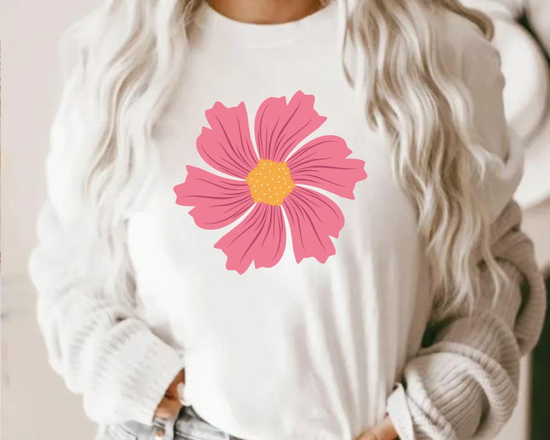 May include: White long-sleeved shirt featuring a large pink flower with a yellow center. The flower design is centered on the shirt's front. The shirt has a crew neck and is made of a soft, comfortable material.