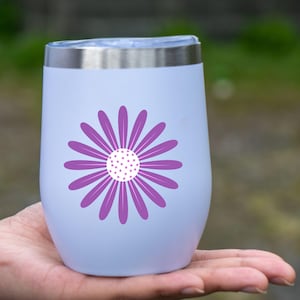 May include: A white insulated wine tumbler with a silver rim and a purple daisy design. The daisy has a white center with purple dots and purple petals. The tumbler is held in a person's hand.