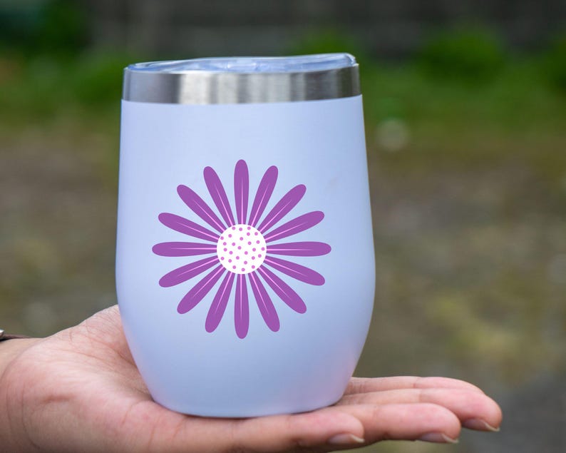 May include: A white insulated wine tumbler with a silver rim and a purple daisy design. The daisy has a white center with purple dots and purple petals. The tumbler is held in a person's hand.