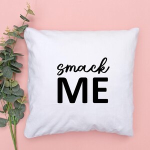 May include: White square pillow with the words "smack ME" printed in black. The word "smack" is in a cursive font, and "ME" is in a bold, block font. The pillow is on a pink background with green foliage.